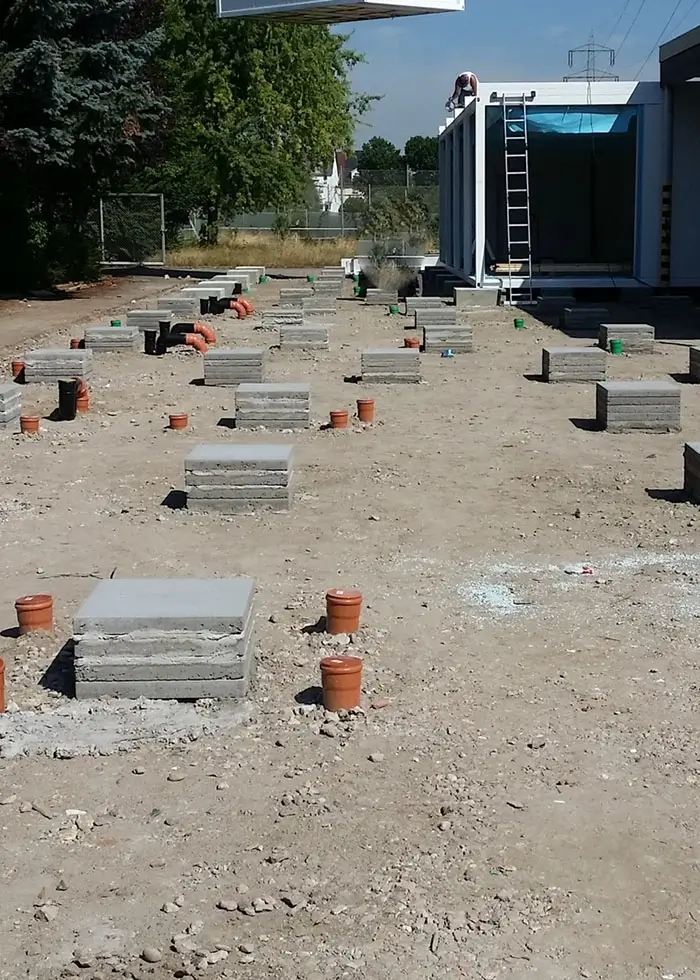 Baugrundstück mit mehreren quadratischen Betonfundamenten und orangefarbenen Rohren in unebenem Erdreich