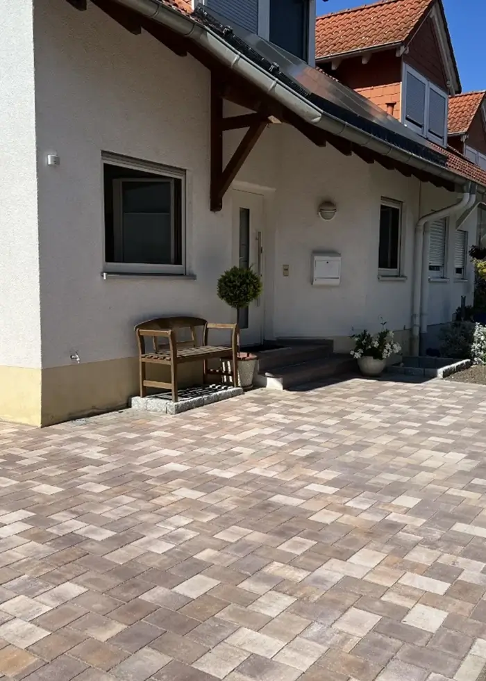 Vorgarten mit gepflastertem Boden, Holzbank, Topfpflanzen und weißem Haus mit Fenstern und Haustür